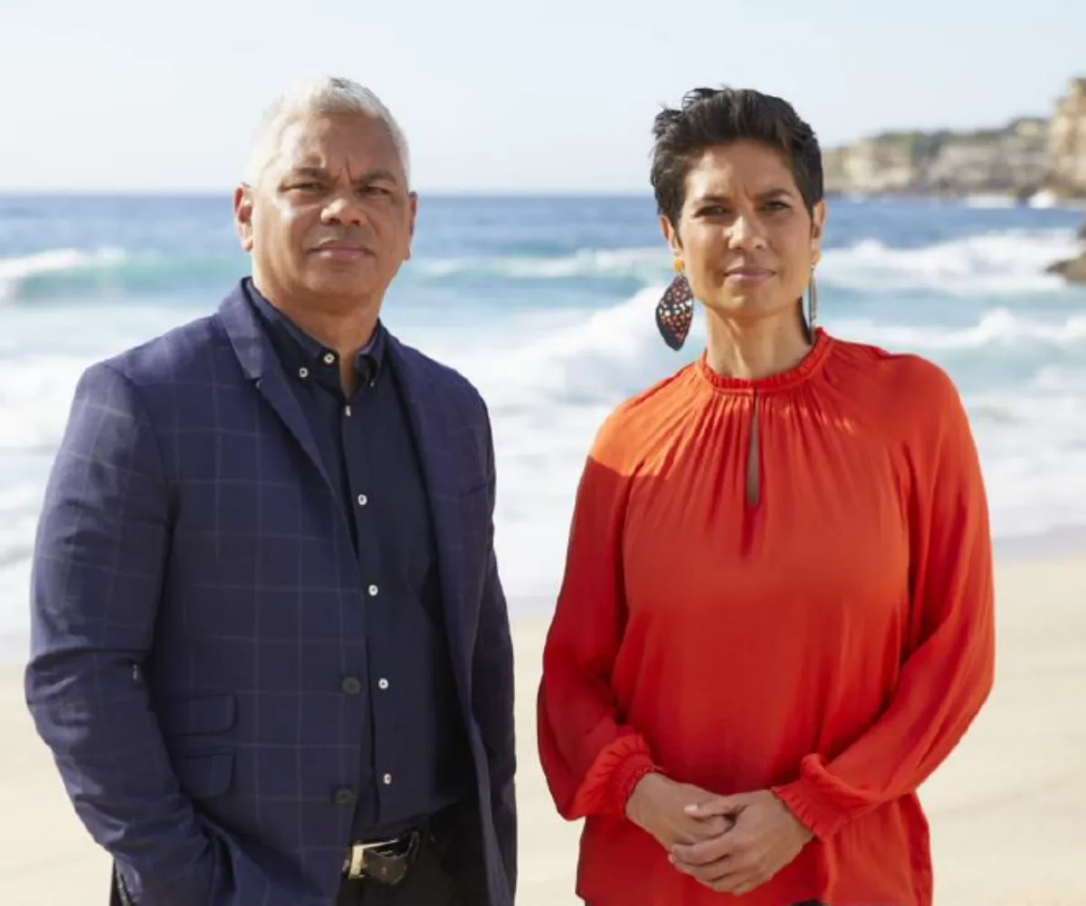 Two people stand on a beach; one in a blue suit, the other in an orange dress. Ocean waves are visible in the background.