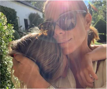 A woman wearing sunglasses hugs a child outdoors, with sunlight streaming through trees in the background.