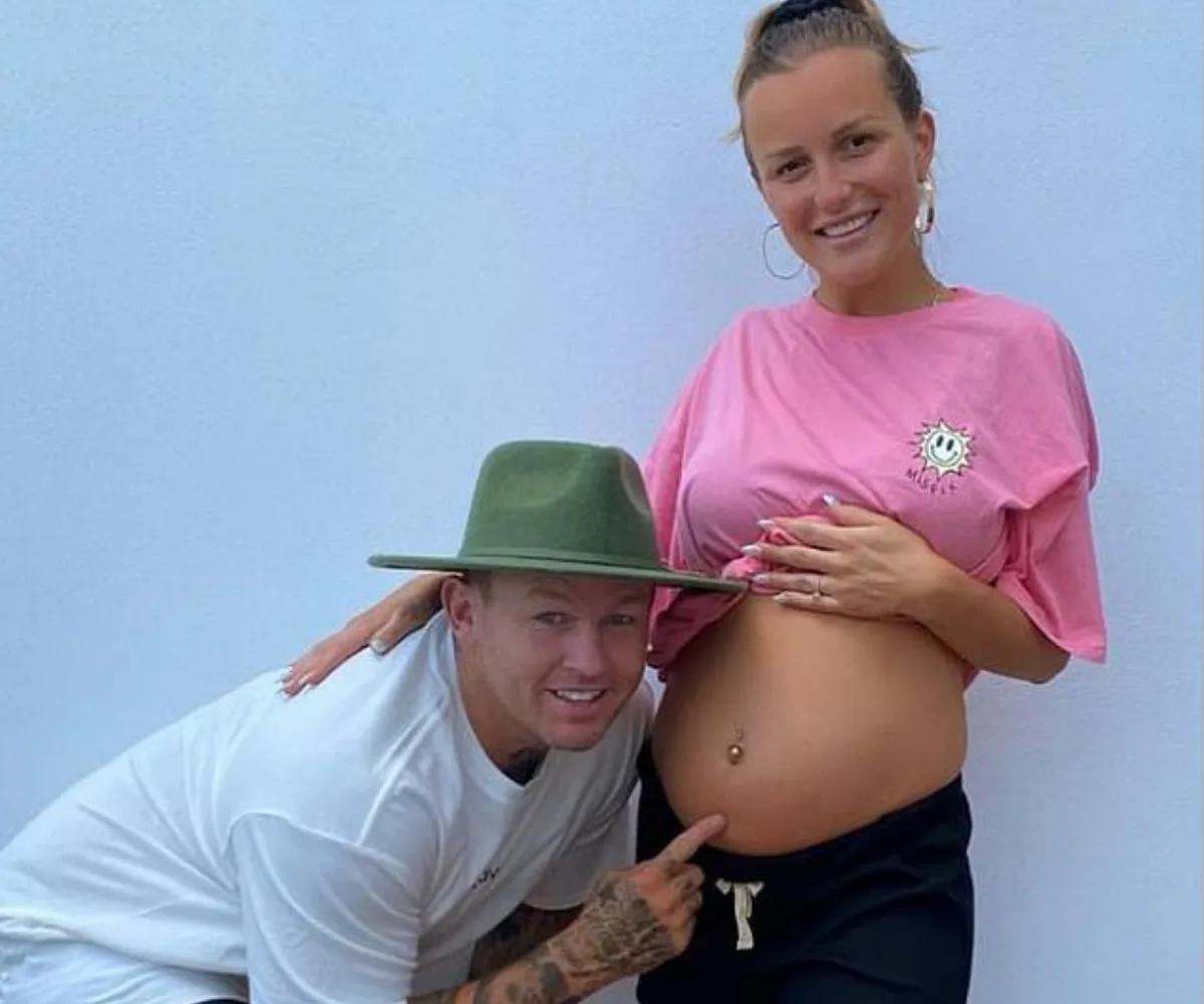 Pregnant woman in pink shirt with smiling man in white, wearing green hat, pointing at her belly.