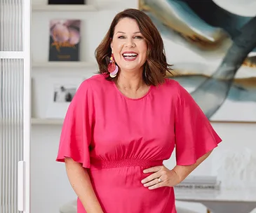 Julia Morris smiling, wearing a bright pink dress, standing in a modern room with abstract artwork and books on the shelf.