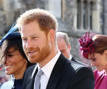 Prince Harry smiling at an outdoor event, accompanied by Meghan Markle and others in formal attire.
