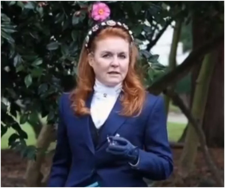 A woman with red hair in a blue outfit stands outdoors, holding an item, with flowers and greenery in the background.