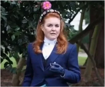 A woman with red hair in a blue outfit stands outdoors, holding an item, with flowers and greenery in the background.