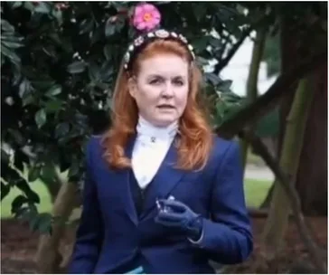 A woman with red hair in a blue outfit stands outdoors, holding an item, with flowers and greenery in the background.