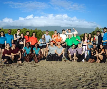 Contestants of The Amazing Race Australia 2021 standing in a group on a sandy beach with a scenic backdrop of trees and mountains.