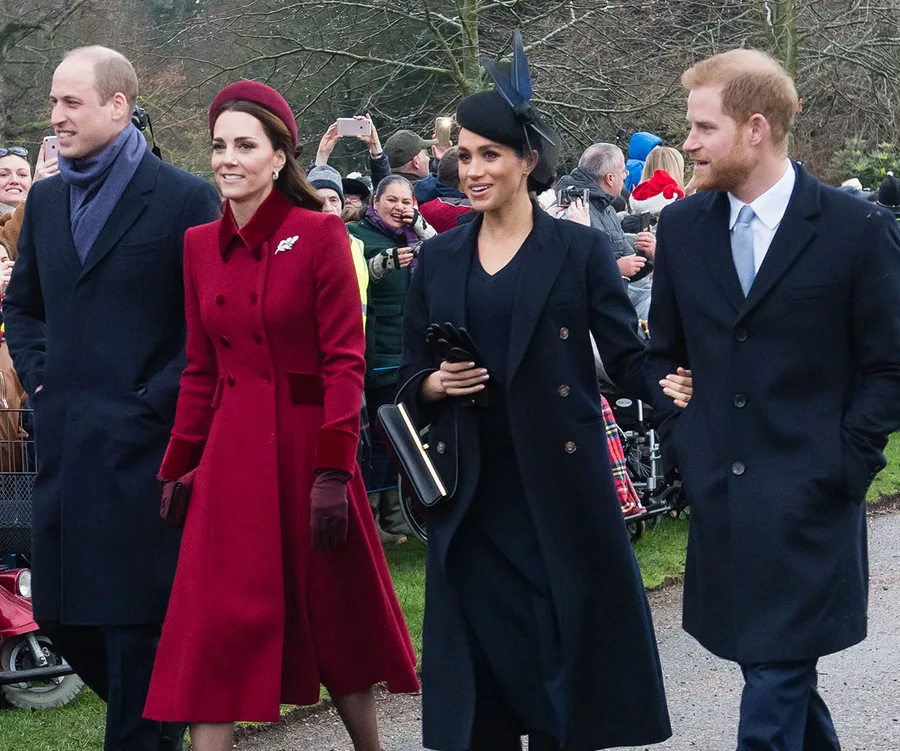 Four individuals in formal winter attire walking outdoors, two are women in red and black coats, two are men in dark coats.