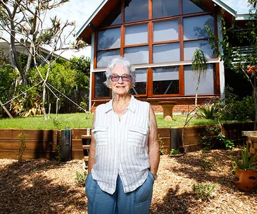 Woman in a white striped shirt standing in front of a modern house with large windows and a garden.