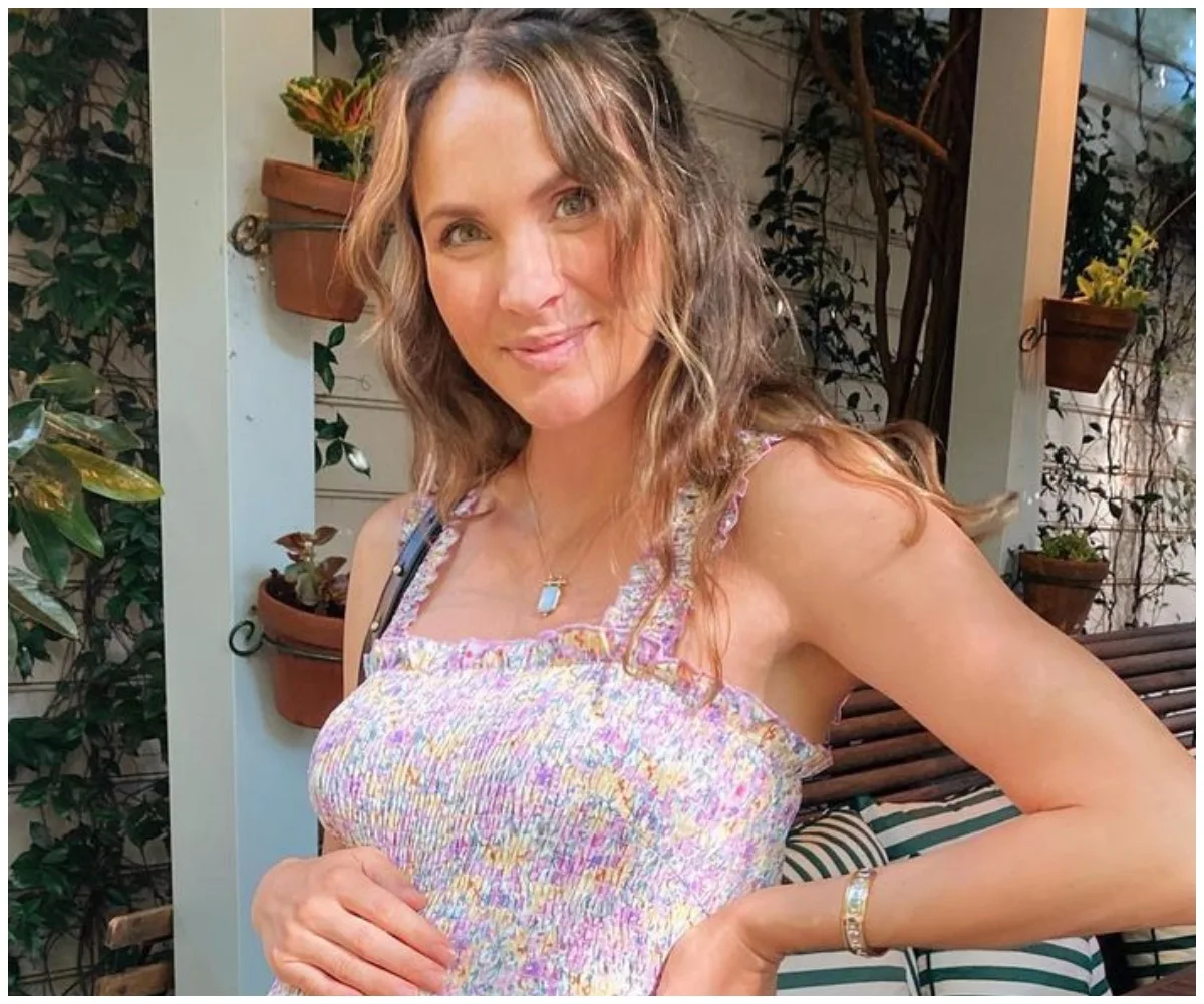 Woman in a floral dress standing outdoors, smiling and cradling her belly, surrounded by plants.