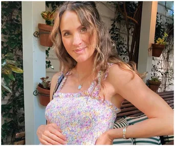 Woman in a floral dress standing outdoors, smiling and cradling her belly, surrounded by plants.