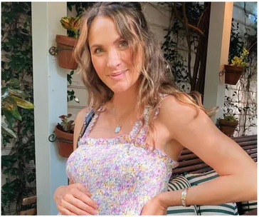 Woman in a floral dress standing outdoors, smiling and cradling her belly, surrounded by plants.