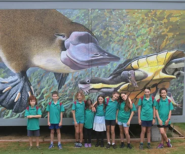 Children in green shirts stand in front of a mural featuring a platypus and a turtle.
