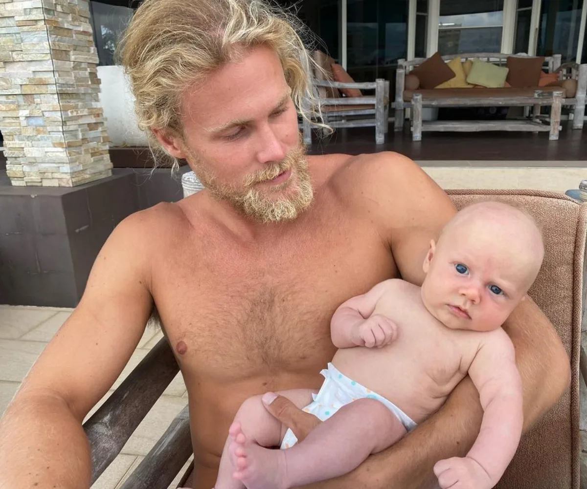 Man with sandy blonde hair holding a baby, sitting on an outdoor patio with rustic furniture in the background.