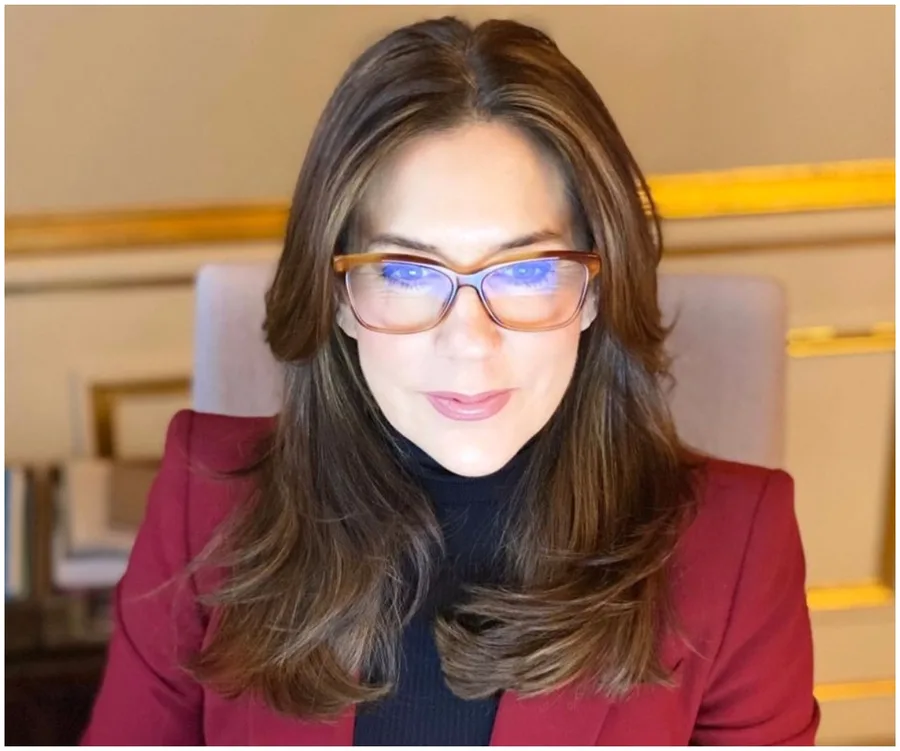 A woman with glasses in a maroon blazer, sitting indoors.