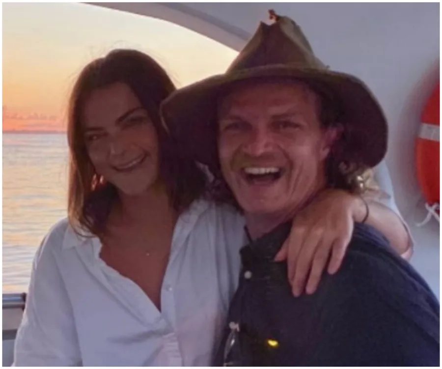 A couple smiling on a boat at sunset with a lifebuoy in the background.