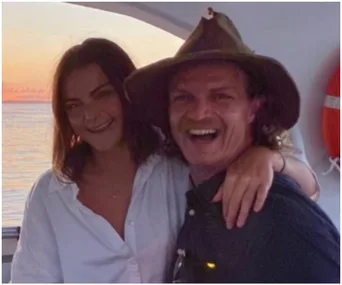 A couple smiling on a boat at sunset with a lifebuoy in the background.
