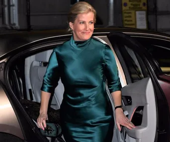 Woman in teal satin dress stepping out of a car, smiling, holding a clutch.