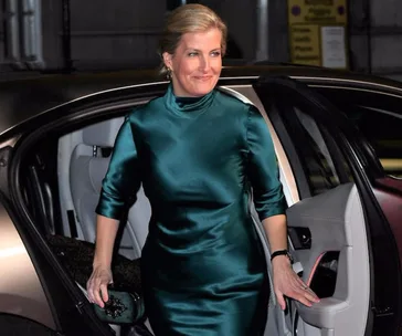 Woman in teal satin dress stepping out of a car, smiling, holding a clutch.