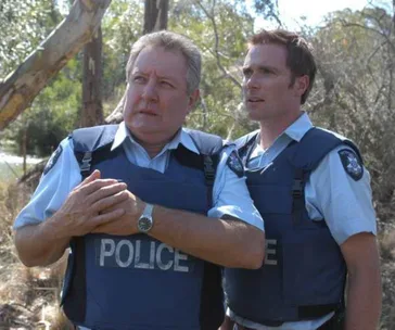 Two police officers in bulletproof vests stand outdoors, appearing concerned, with trees and foliage in the background.