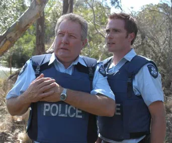 Two police officers in bulletproof vests stand outdoors, appearing concerned, with trees and foliage in the background.