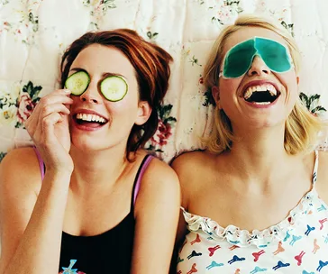 Two women laughing, one with cucumber slices on eyes, the other with a gel eye mask, lying on a floral bedspread.
