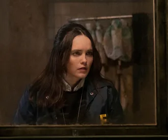 A woman in an FBI jacket looks through a window with concern, standing indoors against a blurred background.
