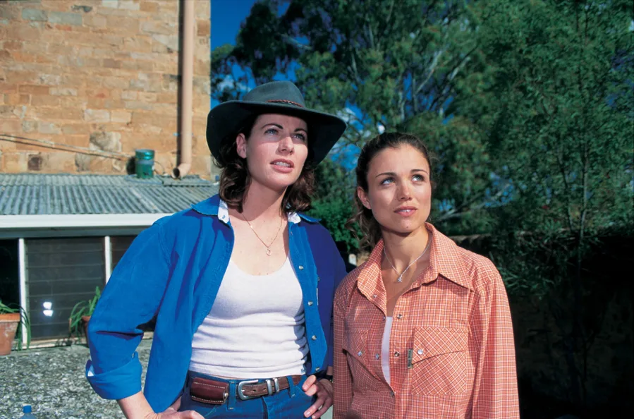 Two women outdoors, one in a blue shirt and hat, the other in a red checkered shirt, gaze into the distance.