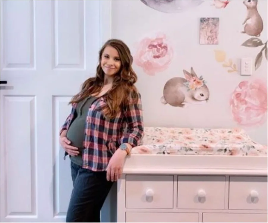 Pregnant woman in plaid shirt poses in nursery with animal wall art and changing table.