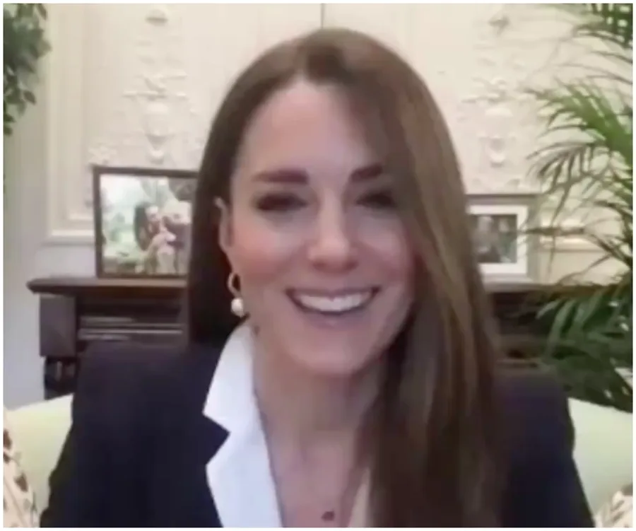 Smiling woman with long hair in a video call, with framed photos and plants in the background.