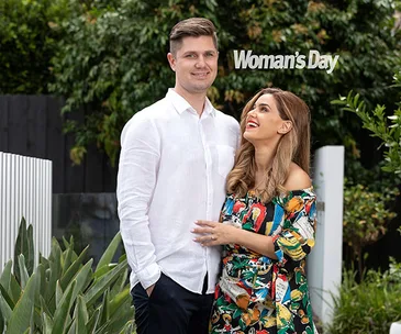 Man in white shirt and woman in colorful dress smiling outdoors, with "Woman's Day" sign in the background.