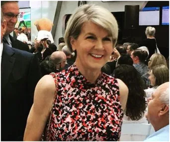 A woman with short hair and a floral dress smiles at an event, surrounded by seated people.