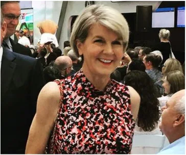 A woman with short hair and a floral dress smiles at an event, surrounded by seated people.