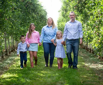 A family of five walks hand-in-hand through a lush orchard, smiling under sunny skies.