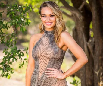 A woman smiles outdoors in a sleeveless, sequined dress with trees in the background.