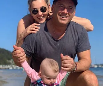Family on a sunny beach day; a man holds a baby's hands while a woman hugs him from behind.