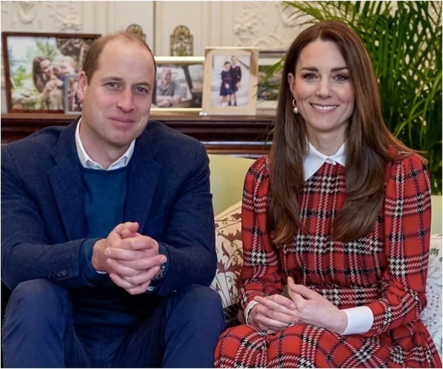 A man and a woman smiling, seated indoors; the woman wears a red tartan dress.