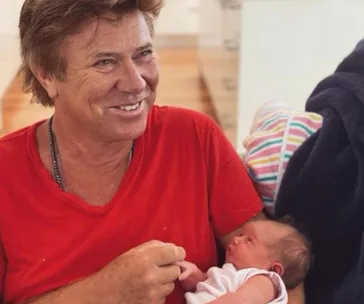 A man in a red shirt smiling while holding a newborn baby wrapped in a white blanket.