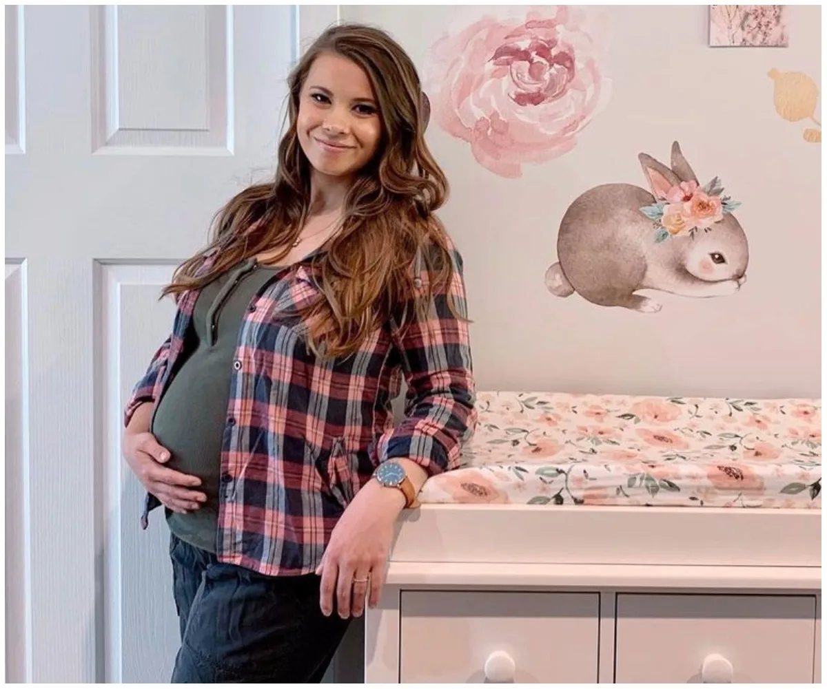 Pregnant woman smiles, holding her belly, standing beside a floral changing table with a bunny wall decal.