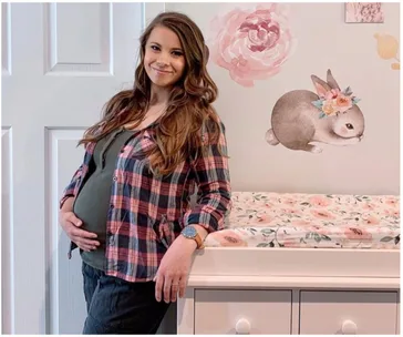 Pregnant woman smiles, holding her belly, standing beside a floral changing table with a bunny wall decal.