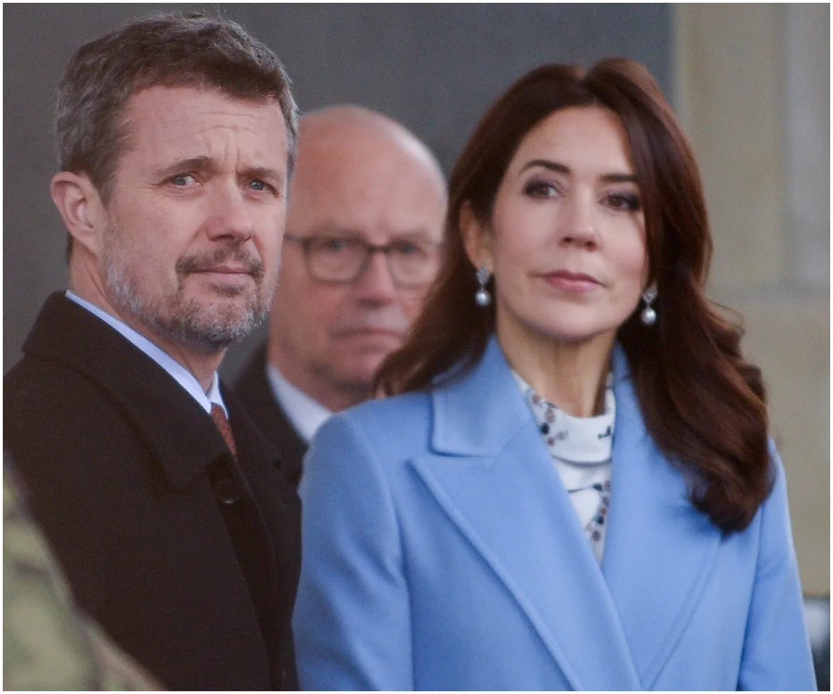 A couple in formal attire stand outdoors, with a man in the background wearing glasses.