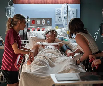 A hospital scene with a patient in bed, surrounded by two concerned women holding his hand and checking on him.