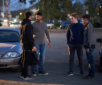 Four men stand near a parked car at dusk, engaged in a tense conversation.