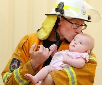 Firefighter in uniform kisses a baby while holding them in his arms, showing tenderness and care.