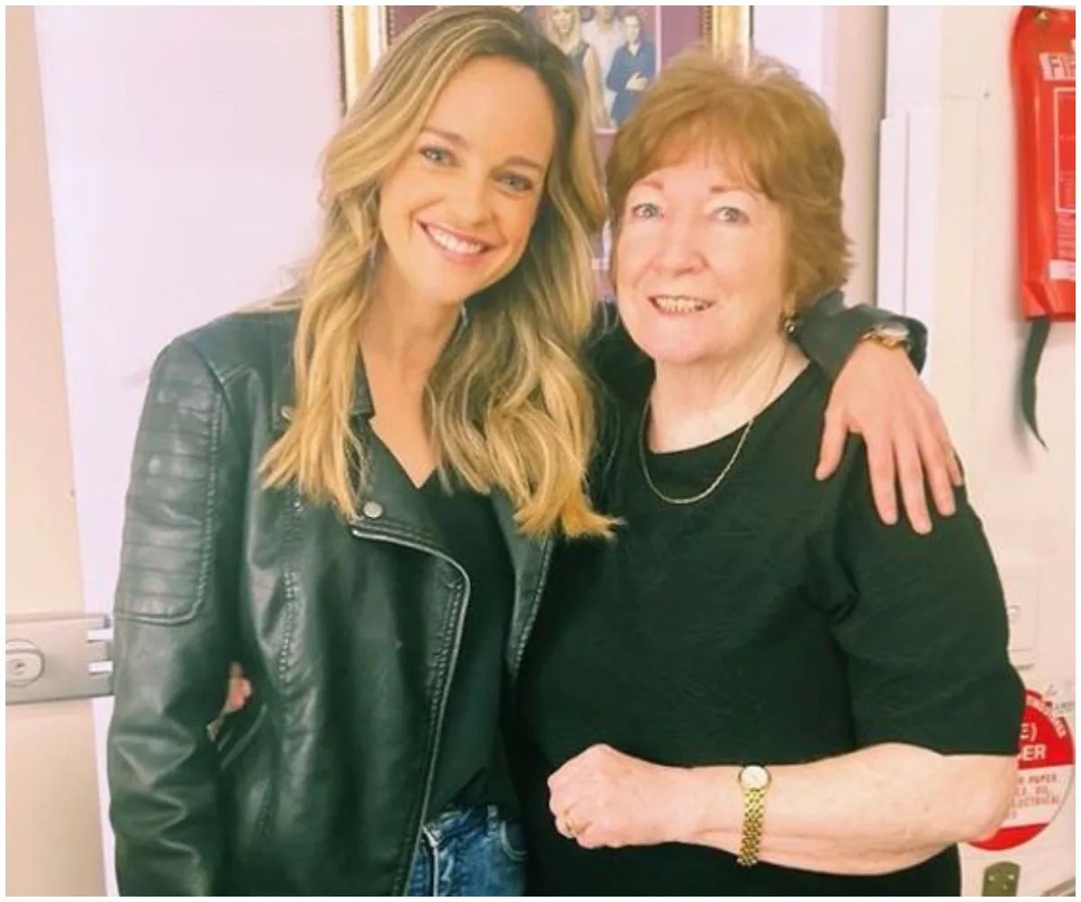 Two women smiling, one in a leather jacket, the other in a black top, standing arm in arm indoors.