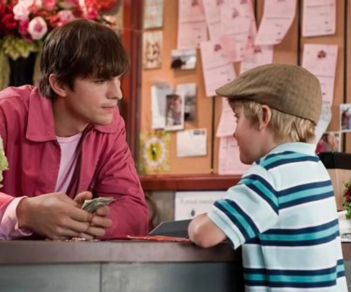 Man in a pink shirt converses with a young boy wearing a striped shirt and cap at a counter with flowers in the background.