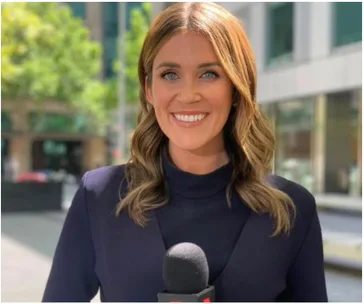 News reporter smiling outside, holding a microphone, with blurred buildings and greenery in the background.