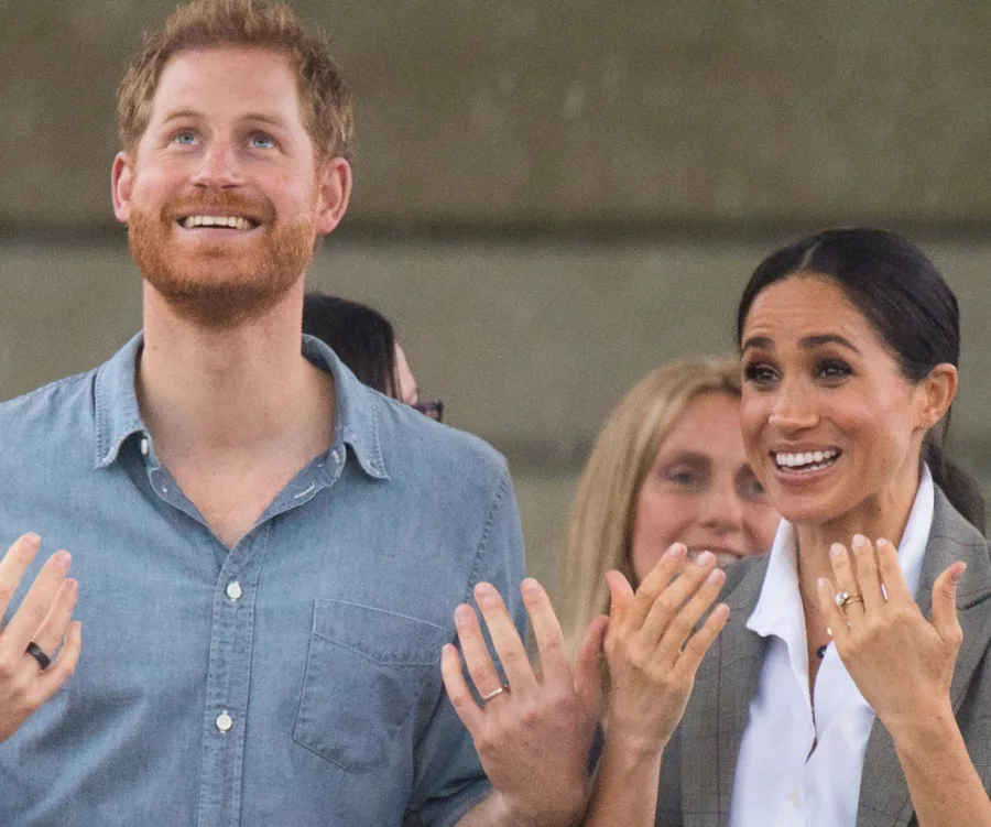Prince Harry and Meghan Markle smiling, looking upward, and clapping at an event in Dubbo.