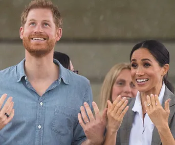 Prince Harry and Meghan Markle smiling, looking upward, and clapping at an event in Dubbo.