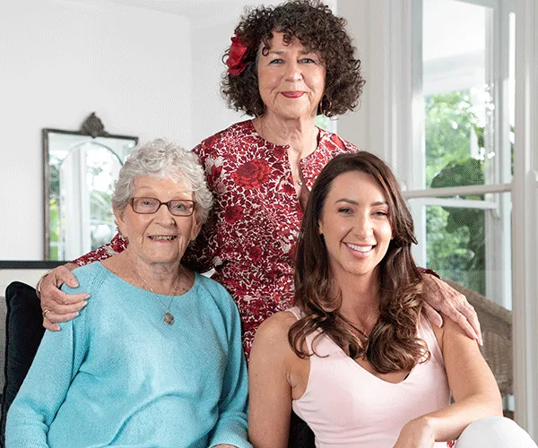 Three women from different generations, smiling together in a cozy living room setting.