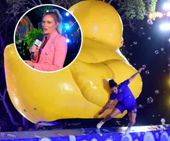 Contestant dodges a giant rubber duck on "Holey Moley" show while host watches with a microphone in hand.