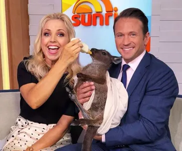 Two people bottle-feed a joey kangaroo on the set of the show "Sunrise", smiling at the camera.